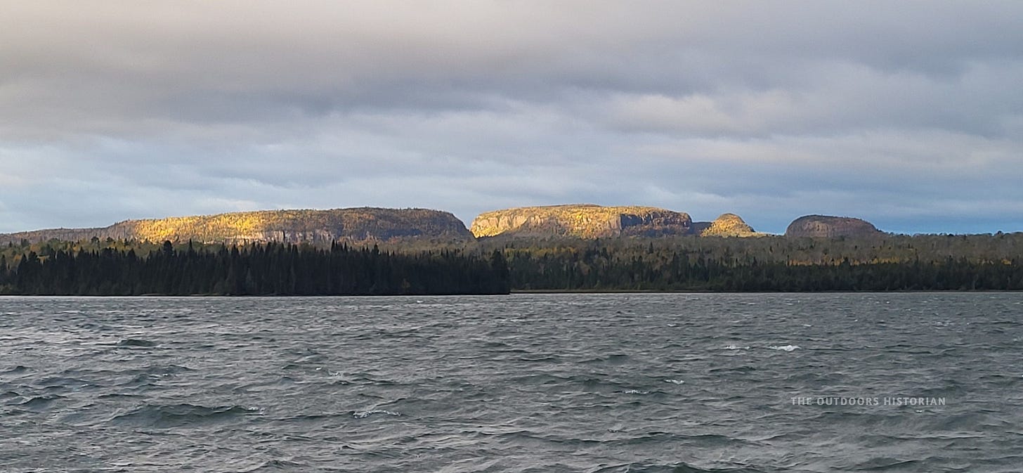 A moody landscape photo. The sky is filled with dark, heavy storm clouds, but a single beam of bright sunlight illuminates the rocky face of the Sleeping Giant.