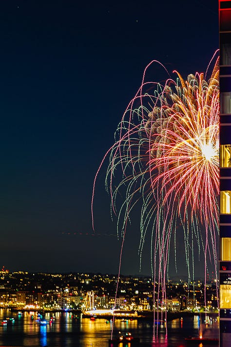 Night fireworks exploding beside Seaport skyscraper with Boston skyline and harbor reflections