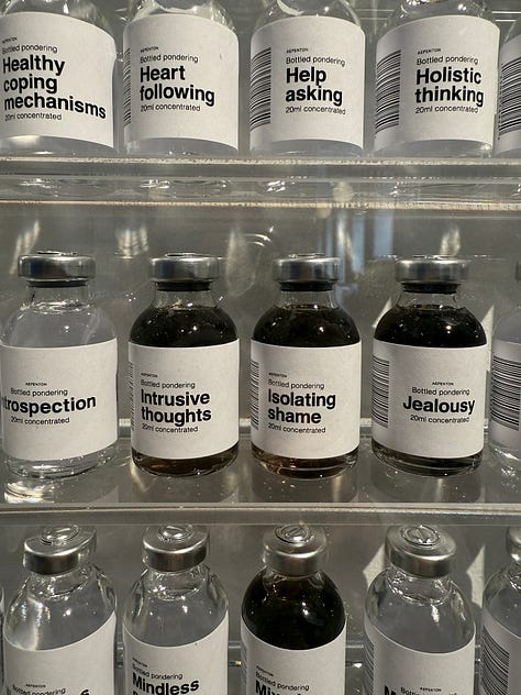 Art installation titled 'Bottled Pondering' by Aepenton, on display at the Design Museum Denmark. The piece features a wall-mounted grid of small glass vials, each labeled with a different abstract or emotional concept, such as 'Inner Critic Softener' or 'Courage Enhancer.' The vials are arranged in neat rows within a transparent display case, evoking the look of a scientific archive or apothecary cabinet of thoughts and feelings.