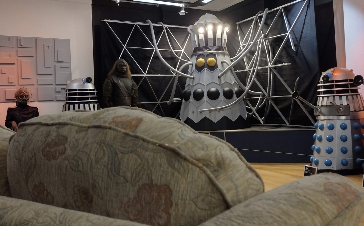 Two Daleks, an Ogron, Davros and the Dalek Emperor, with the back of a sofa in the foreground.
