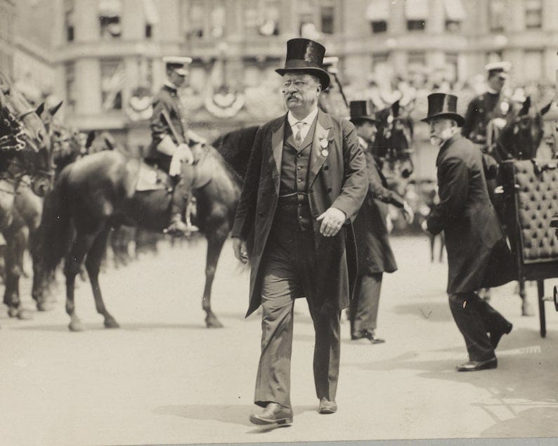 May include: A man in a top hat and suit walks down a street, surrounded by people and horses. He is likely a politician or other important figure.