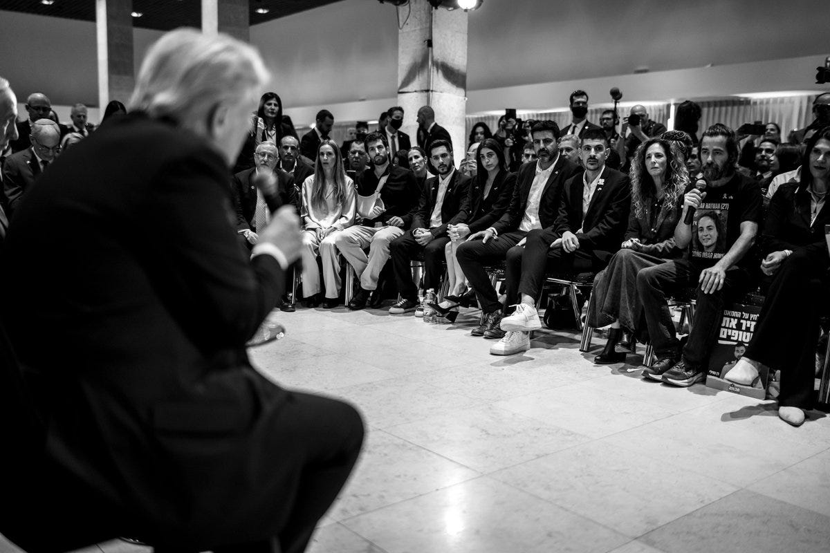 Black-and-white photograph of a diverse group of people seated and standing in a semi-circle on a tiled floor in what appears to be an indoor venue with pillars and overhead lighting. Several individuals wear formal attire including suits and dresses, with some seated on chairs and others standing or gesturing. The group includes men and women of various ages and ethnicities, focused toward a central area.