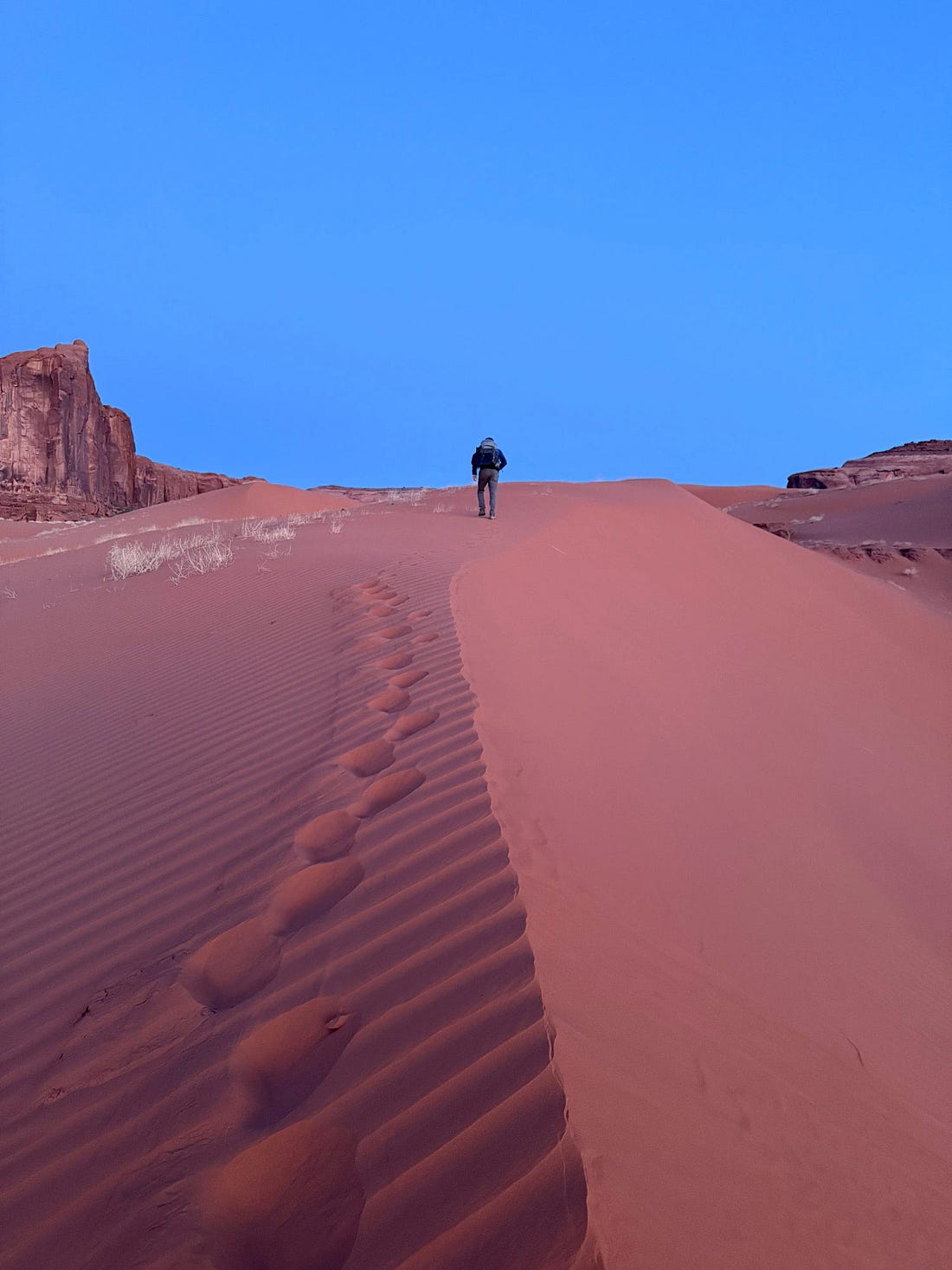 Hiker climbing the Sand Springs dunes at dawn, overlooking sandstone monoliths where small groups of Ancestral Puebloans once built cliff dwellings and granaries.