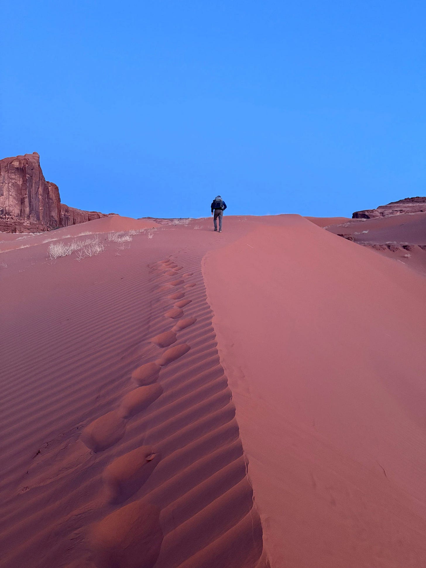 Hiker climbing the Sand Springs dunes at dawn, overlooking sandstone monoliths where small groups of Ancestral Puebloans once built cliff dwellings and granaries.