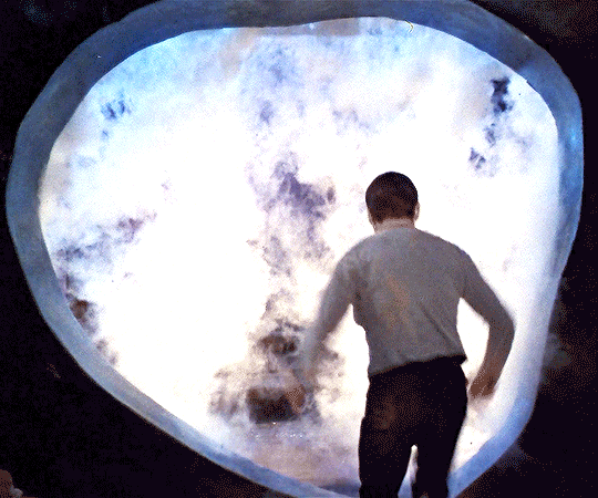 Gif da Star Trek. Un uomo vestito con una camicia chiara e pantaloni scuri, visto da dietro, si protende in avanti verso un portale luminoso e ovale. Il portale emana una luce bianca e bluastra intensa con un effetto che ricorda fumo, nuvole o un campo di energia turbolento al suo interno, in netto contrasto con l'ambiente circostante che è scuro.