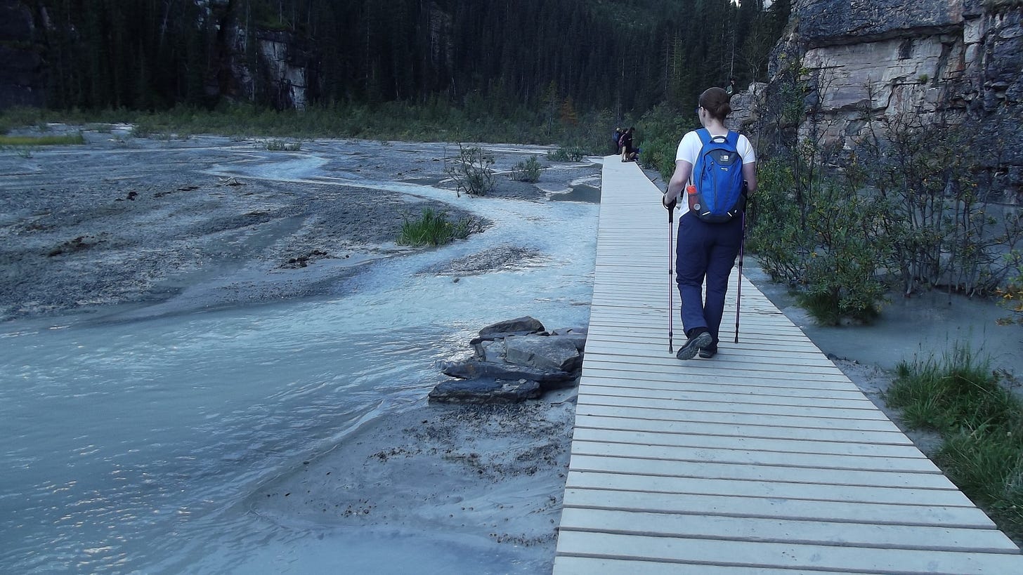 Lisa is walking away from the camera on a wooden bridge over a salt plain, she is resting on two walking sticks and carries a rucksack on her back