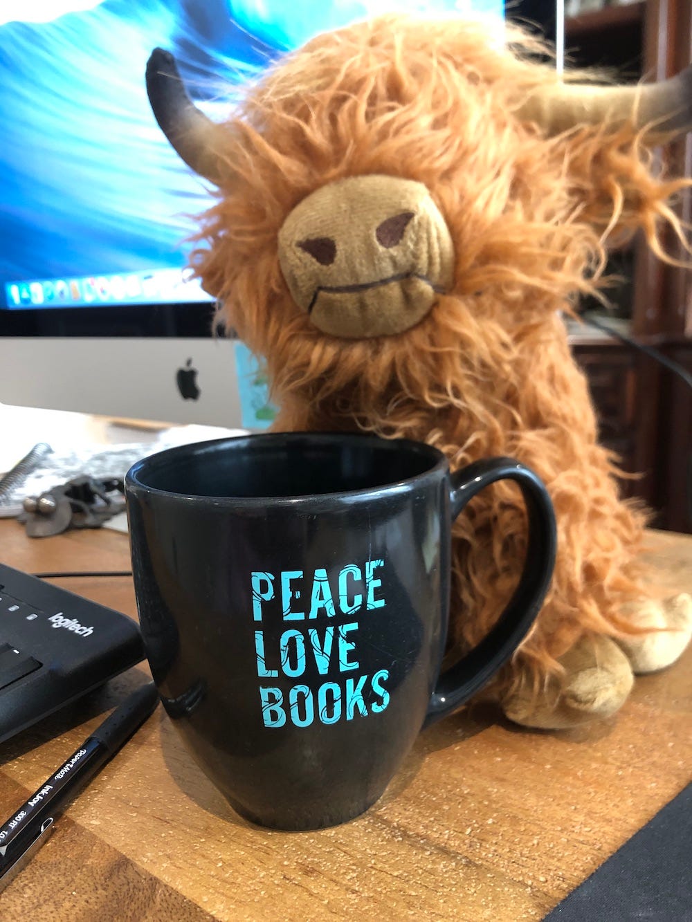 A scruffy stuffed highland cow on a computer desk with a coffee cup that reads "Peace, Love, Books"