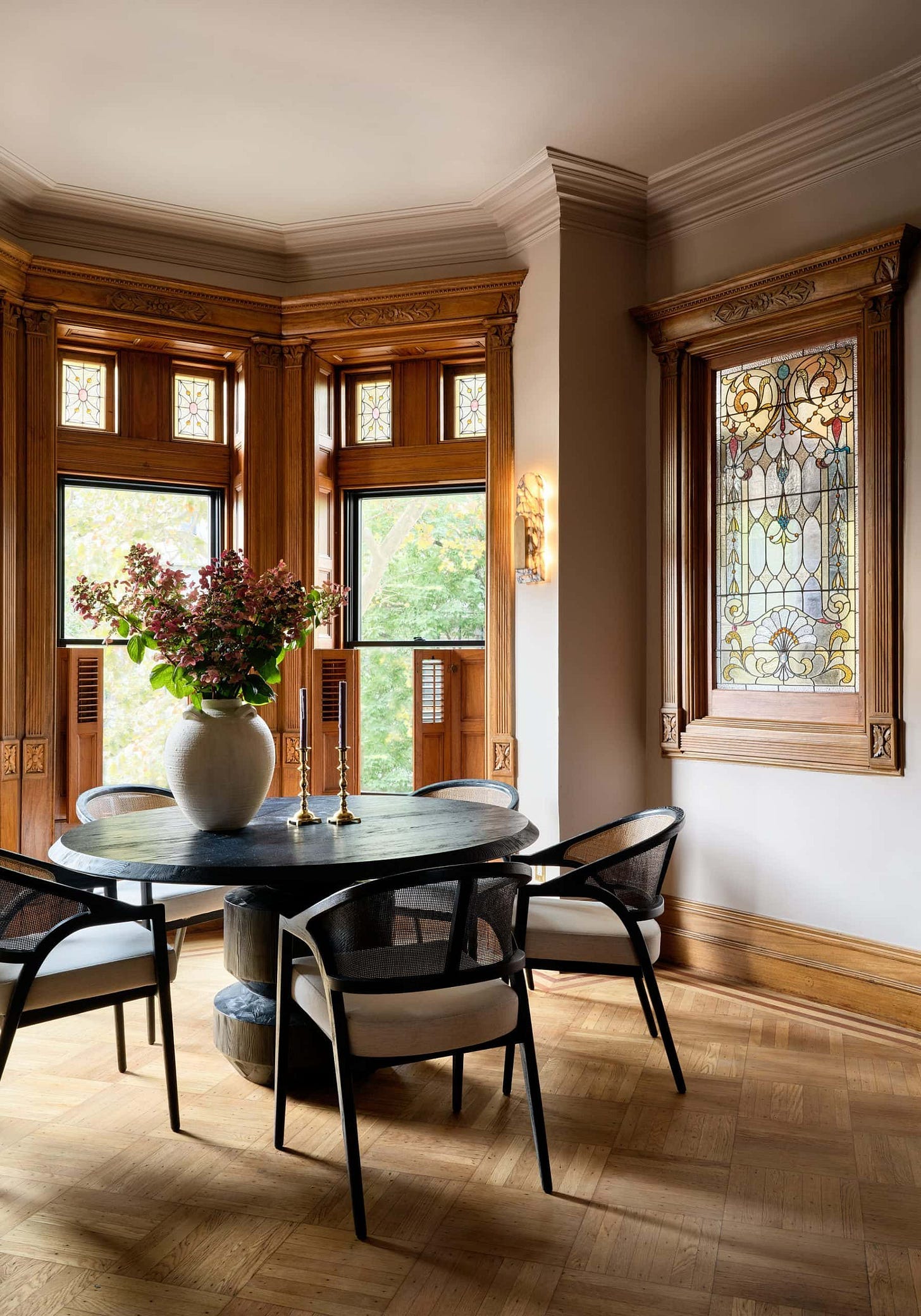 A round black table with four chairs is set in a dining area featuring wooden window frames, stained glass, and a large vase with flowers.