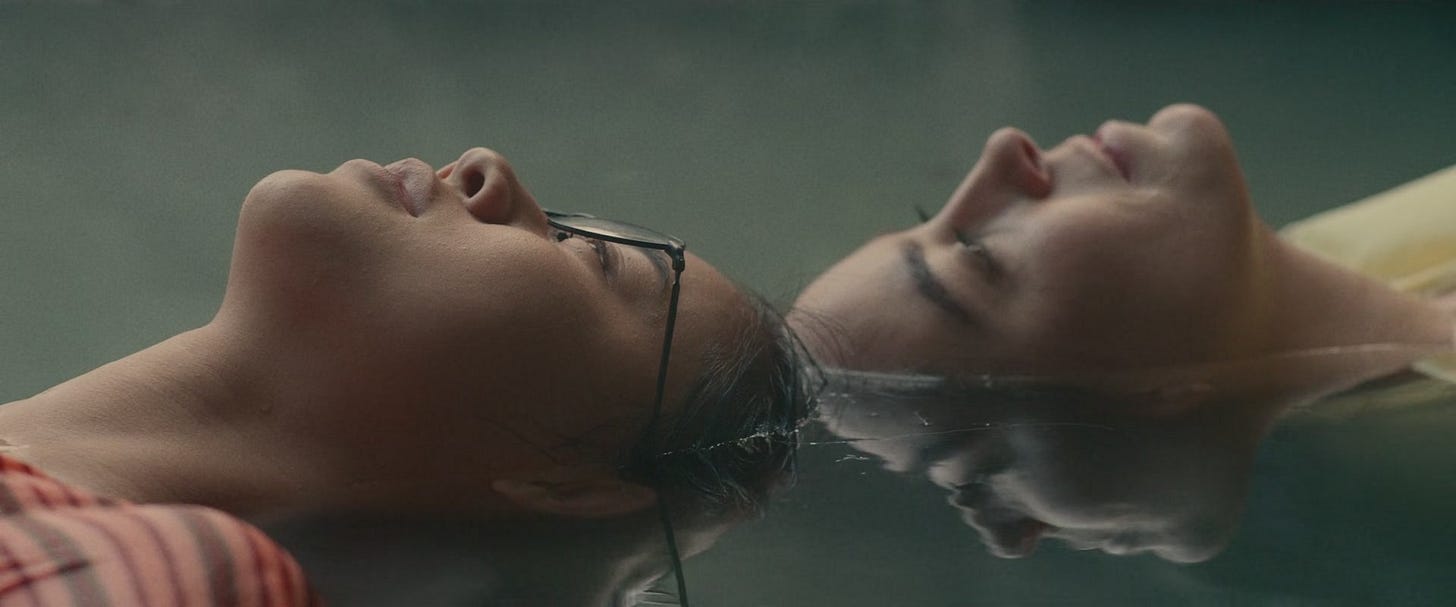 A still from the film 'The Hal of It'. Ellie and Aster lie in the hotspring with their faces barely touching. Ellie wears glasses and a red shirt. Their faces are reflected in the water. A still from the film 'The Hal of It'. Ellie and Aster lie in the hotspring with their faces barely touching. Ellie wears glasses and a red shirt. Their faces are reflected in the water.