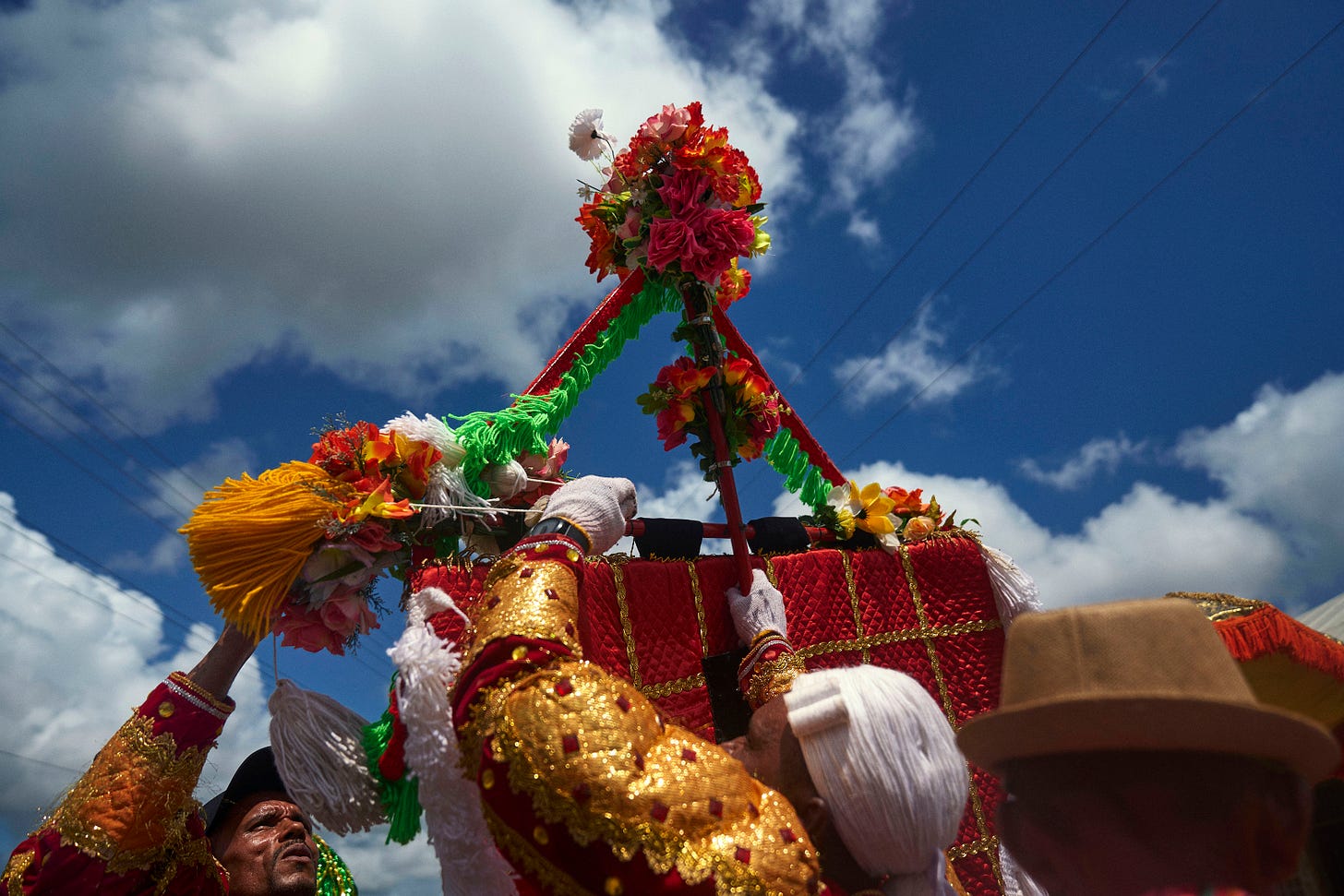 People dressed in colorful, traditional attire hold up a decorated structure with flowers and bright tassels under a blue sky with clouds, participating in a festive outdoor celebration.