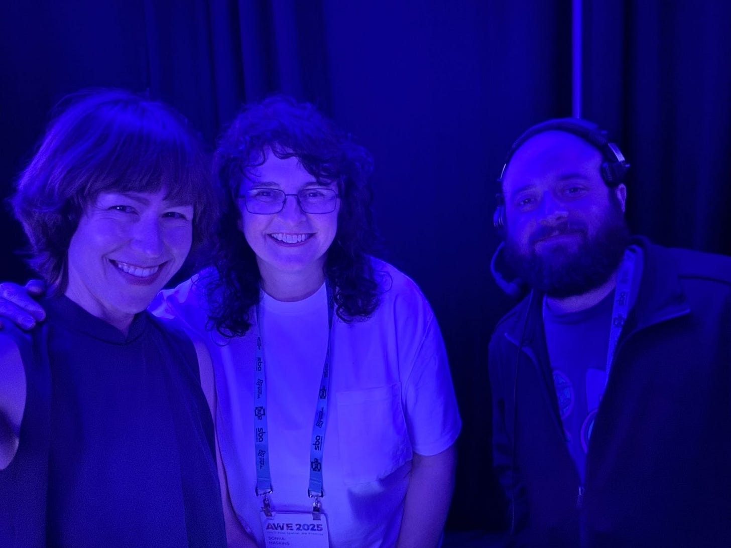 Three people smiling under blue stage lighting at AWE 2025. The person on the left is wearing a sleeveless top, the person in the middle has curly hair, glasses, and an AWE 2025 badge, and the person on the right is wearing a headset. Three people smiling under blue stage lighting at AWE 2025. The person on the left is wearing a sleeveless top, the person in the middle has curly hair, glasses, and an AWE 2025 badge, and the person on the right is wearing a headset.