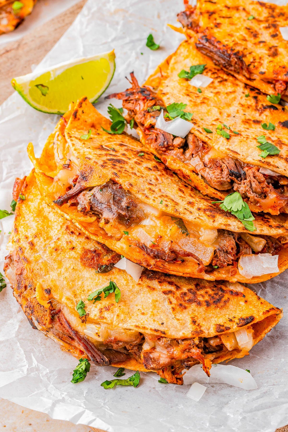Three folded birria tacos filled with meat and melted cheese, garnished with chopped onions and cilantro, served with a lime wedge on parchment paper.