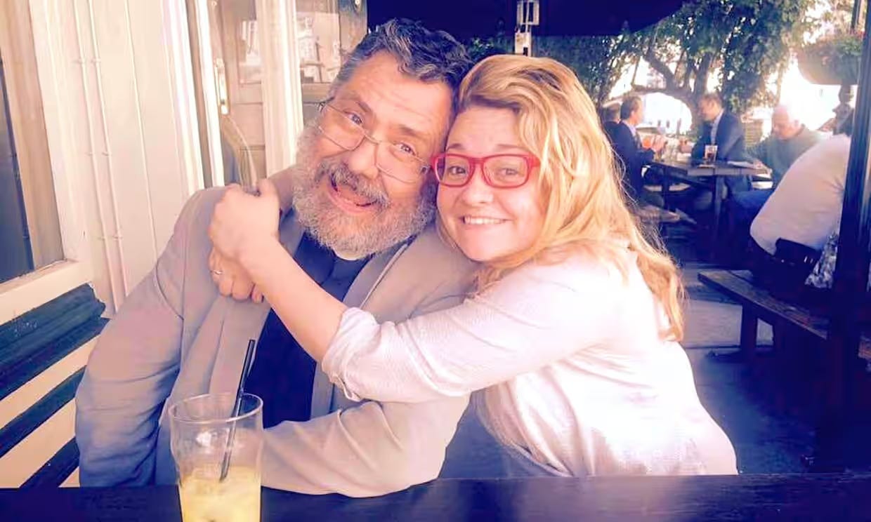 Hannah, younger and blonde with red glasses, hugs her dad, a bearded man wearing a clerical collar.