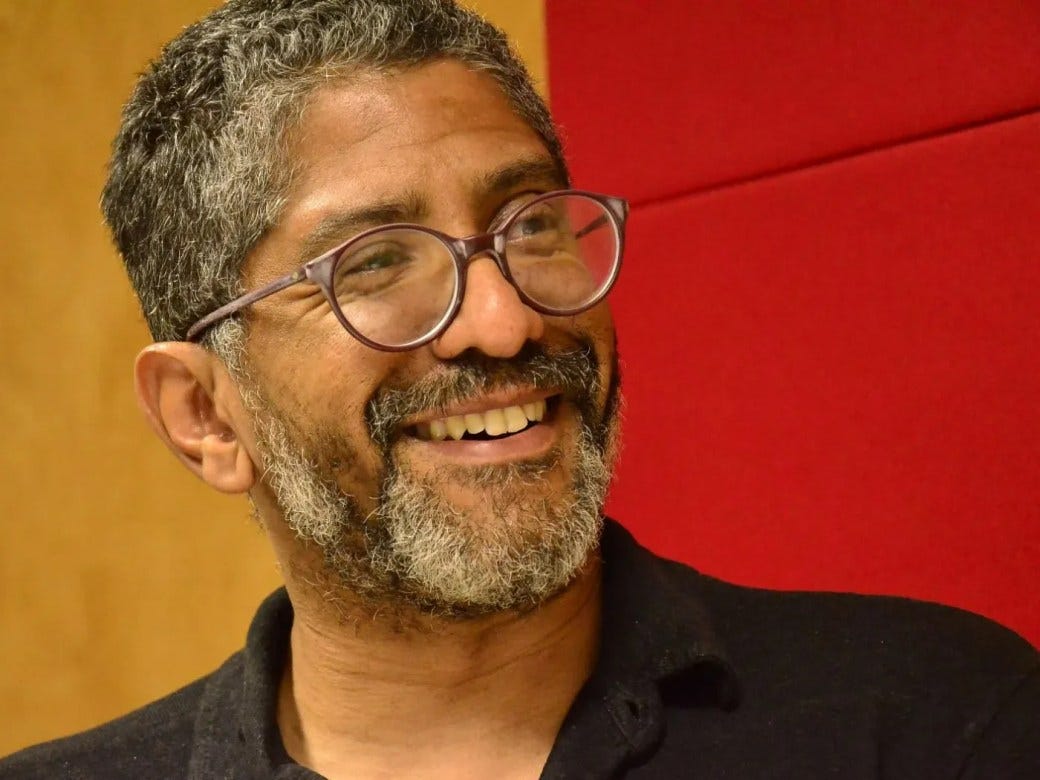 #descrevemospravocê  Fotografia de Jeferson Tenório: um homem sorridente em ambiente interno. Ele tem cabelo curto e grisalho, usa barba e bigode também grisalhos. Veste uma camisa preta com gola polo e está de óculos de armação redonda e transparente, com lentes grandes. O homem olha para o lado, com expressão alegre e descontraída, revelando os dentes ao sorrir. Ao fundo, parte da parede é revestida por painéis vermelhos, enquanto outra parte é de madeira clara, compondo um cenário acolhedor e moderno. A luz está suave e destaca bem os traços do rosto.