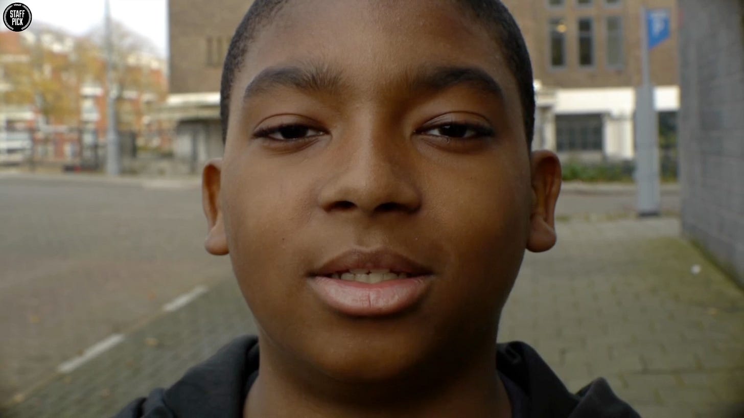 Close-up portrait of a Black teenage boy with a gentle smile, wearing a dark hoodie, photographed outdoors with buildings in the background.