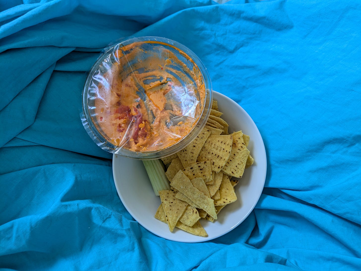 ID. A top down photo of an almost finished pot of roasted red pepper hummus and a bowl of corn chips on top of a bright blue sheet. ID. A top down photo of an almost finished pot of roasted red pepper hummus and a bowl of corn chips on top of a bright blue sheet.