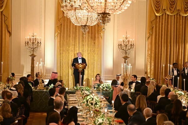 President Trump, wearing a bow tie and a black suit, stands in a ornately decorated room in front of long tables of seated people.