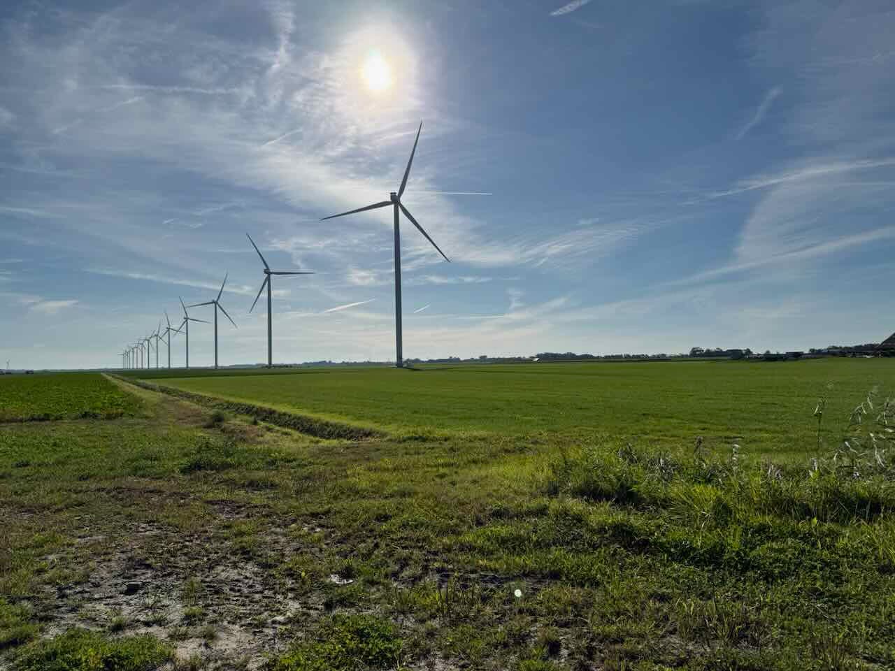 A row of tall wind turbines stands in a green, open field under a bright blue sky with wispy clouds in North Holland. The sun shines above, casting light across the flat landscape that stretches toward the horizon.