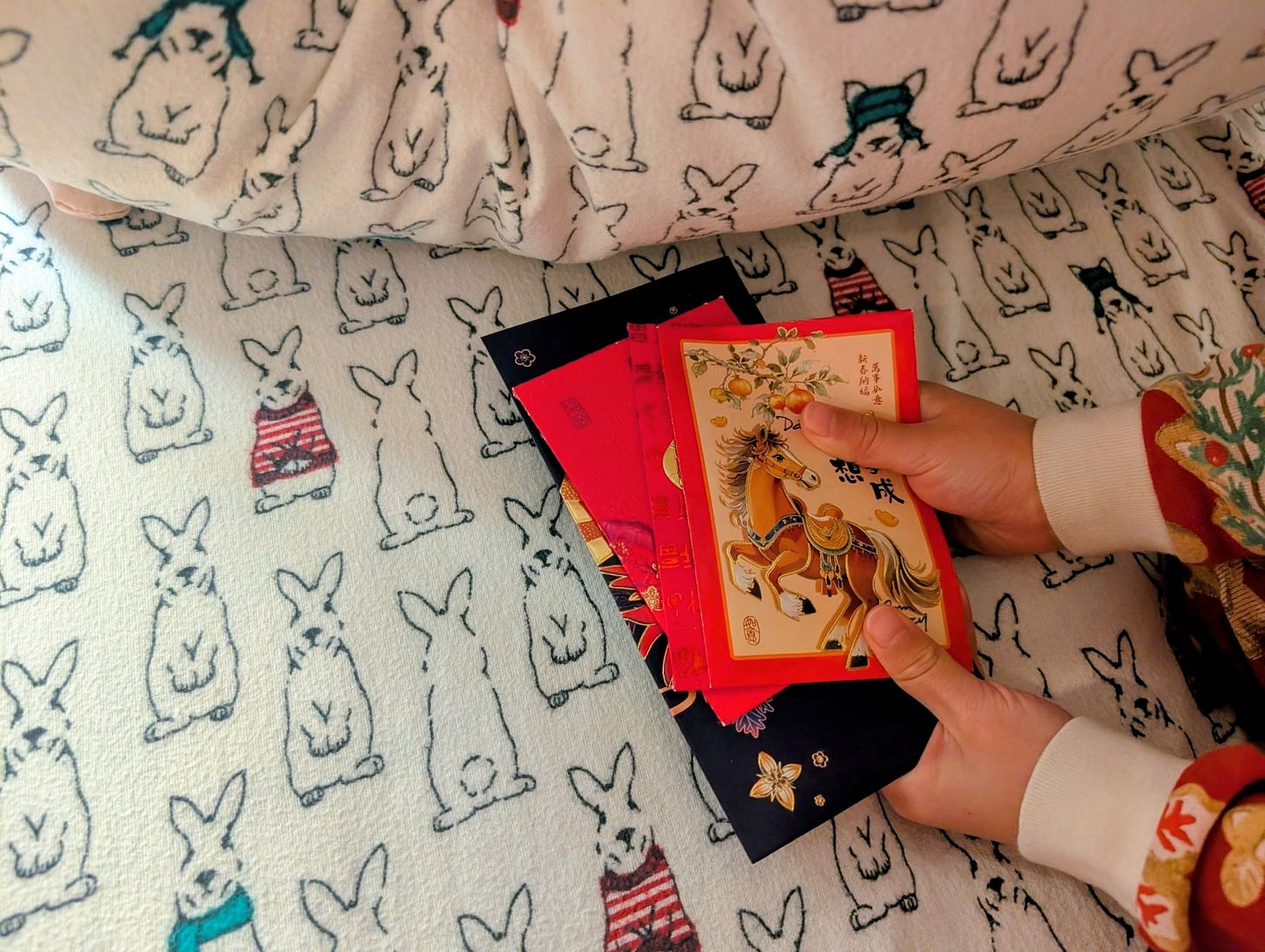 Little hands hold a stack of red envelopes under a pillow. The pillow and sheets have a rabbit pattern.