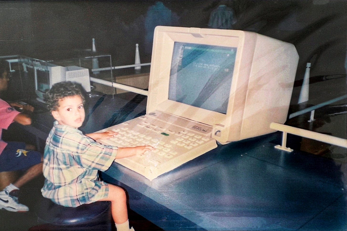A young child in a plaid shirt sits at a large, vintage computer terminal with a dark screen at an exhibit in the 1990s. A young child in a plaid shirt sits at a large, vintage computer terminal with a dark screen at an exhibit in the 1990s.