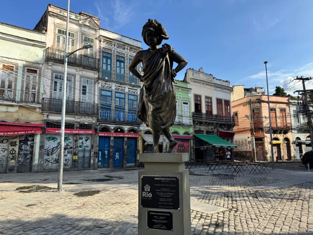 #descrevemospravocê  A imagem mostra uma praça de calçamento em pedras, com destaque para uma estátua de bronze em primeiro plano. A escultura representa uma mulher negra em pose altiva, com uma das pernas levemente à frente, mãos na cintura, olhar erguido e expressão determinada. Ela veste saia rodada e turbante na cabeça, remetendo a vestimentas tradicionais afro-brasileiras. A figura está sobre um pedestal retangular de pedra, que contém uma placa metálica identificando a homenageada: Mercedes Baptista, importante bailarina, coreógrafa e pioneira na valorização da cultura afro-brasileira na dança.  Ao fundo, há um conjunto de prédios antigos e coloridos com fachadas em estilo colonial e eclético. As construções apresentam varandas com grades de ferro e portas e janelas altas. Algumas fachadas estão degradadas e cobertas por pichações e grafites. Toldos vermelhos e verdes indicam o uso comercial dos imóveis térreos, apesar de alguns estarem aparentemente fechados. Em frente às lojas, há cadeiras e mesas dobráveis, sugerindo a presença de bares ou cafés na região.  O céu está claro e azul, e a iluminação solar reforça os detalhes da escultura e das construções. A cena foi registrada durante o dia e transmite a atmosfera histórica e cultural da área urbana retratada, provavelmente no centro do Rio de Janeiro, onde Mercedes Baptista fez história.
