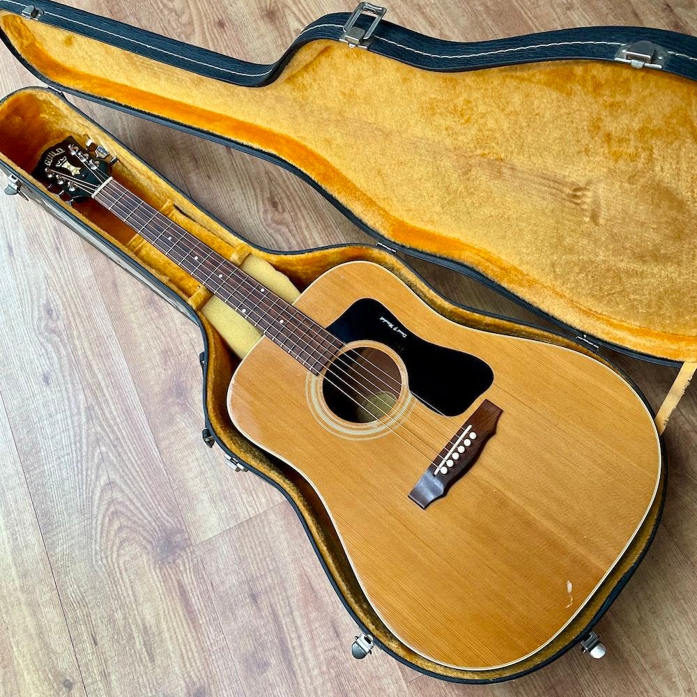 n open guitar case on a polished wooden floor, holding a 1973 Guild D‑40 with a small foam block under the neck and the owner’s name faintly visible on the scratchplate. n open guitar case on a polished wooden floor, holding a 1973 Guild D‑40 with a small foam block under the neck and the owner’s name faintly visible on the scratchplate.