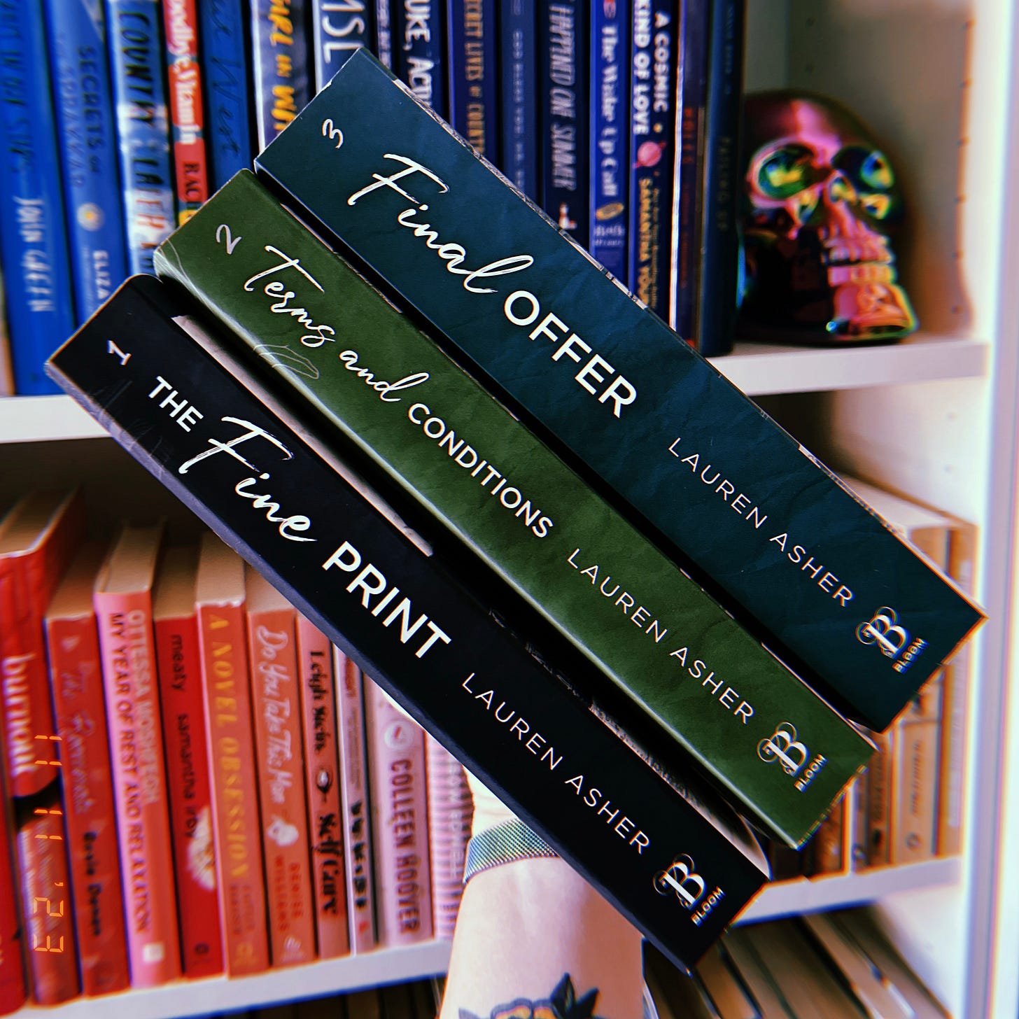 A hand holding a stack of books in front of a bookshelf. The books are The Fine Print, Terms and Conditions, and Final Offer, all by Lauren Asher. A hand holding a stack of books in front of a bookshelf. The books are The Fine Print, Terms and Conditions, and Final Offer, all by Lauren Asher.