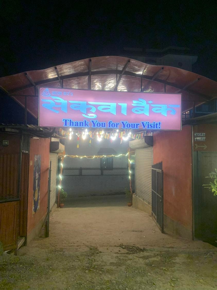 A well-lit archway over a stone path leads to a main road. Brilliantly illuminated above is a sign in Nepali, with the English words, “Thank you for your visit” inscribed beneath.