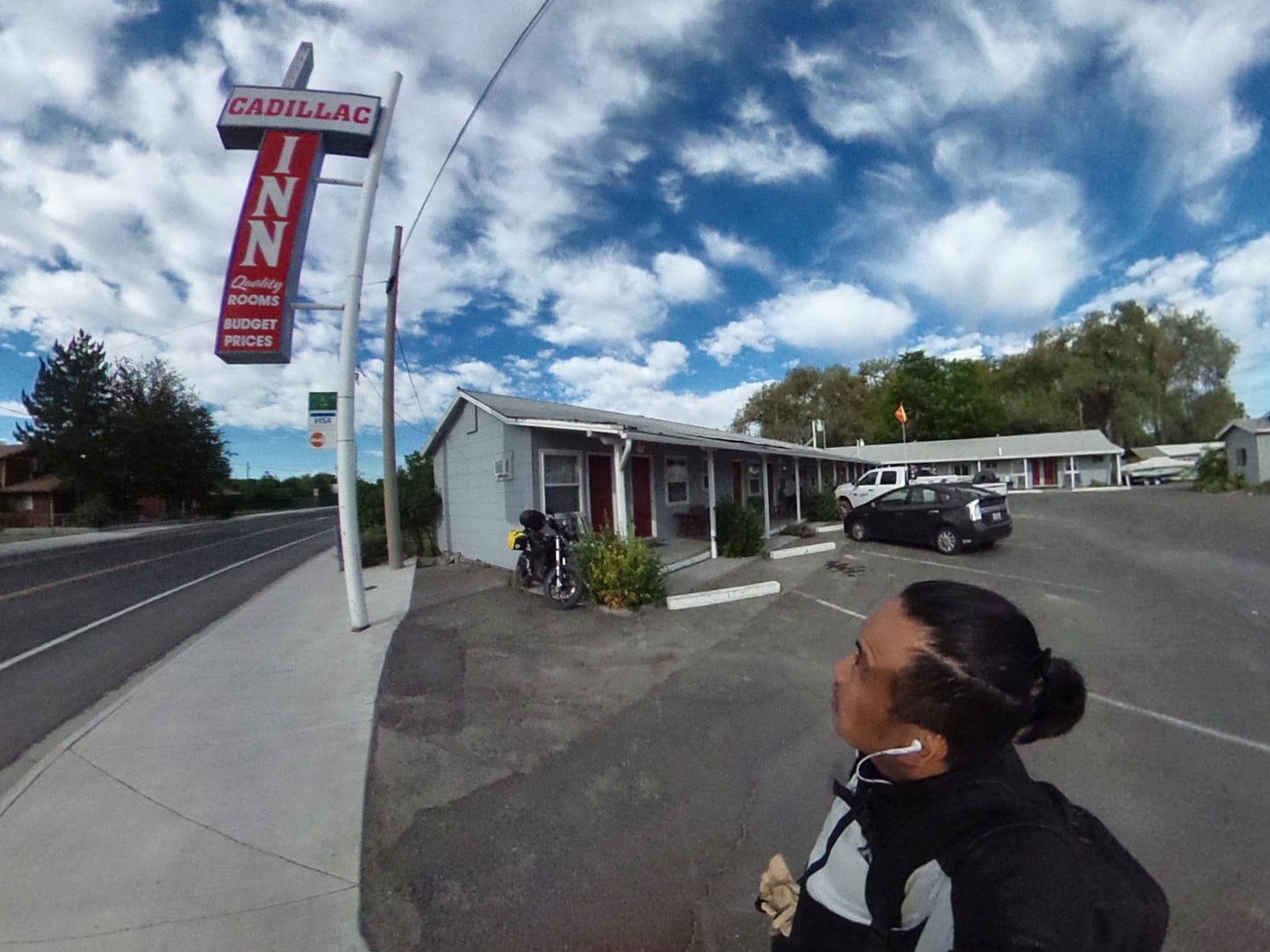 Distorted photo of the author standing in a motel parking courtyard. He is wearing motorcycle riding gear, earbuds, and a man bun looking up at old roadside "Cadillac Inn" sign. "Quality ROOMS. BUDGET PRICES" The motel can be seen in the background, and outside the last room in the motel sits a black electric motorcycle on its sidestand. There is a black Toyota Prius and white, 4-door pickup truck in the courtyard. Green trees dot the horizon. The adjacent road has no cars. The sky is blue with large, puffy clouds.
