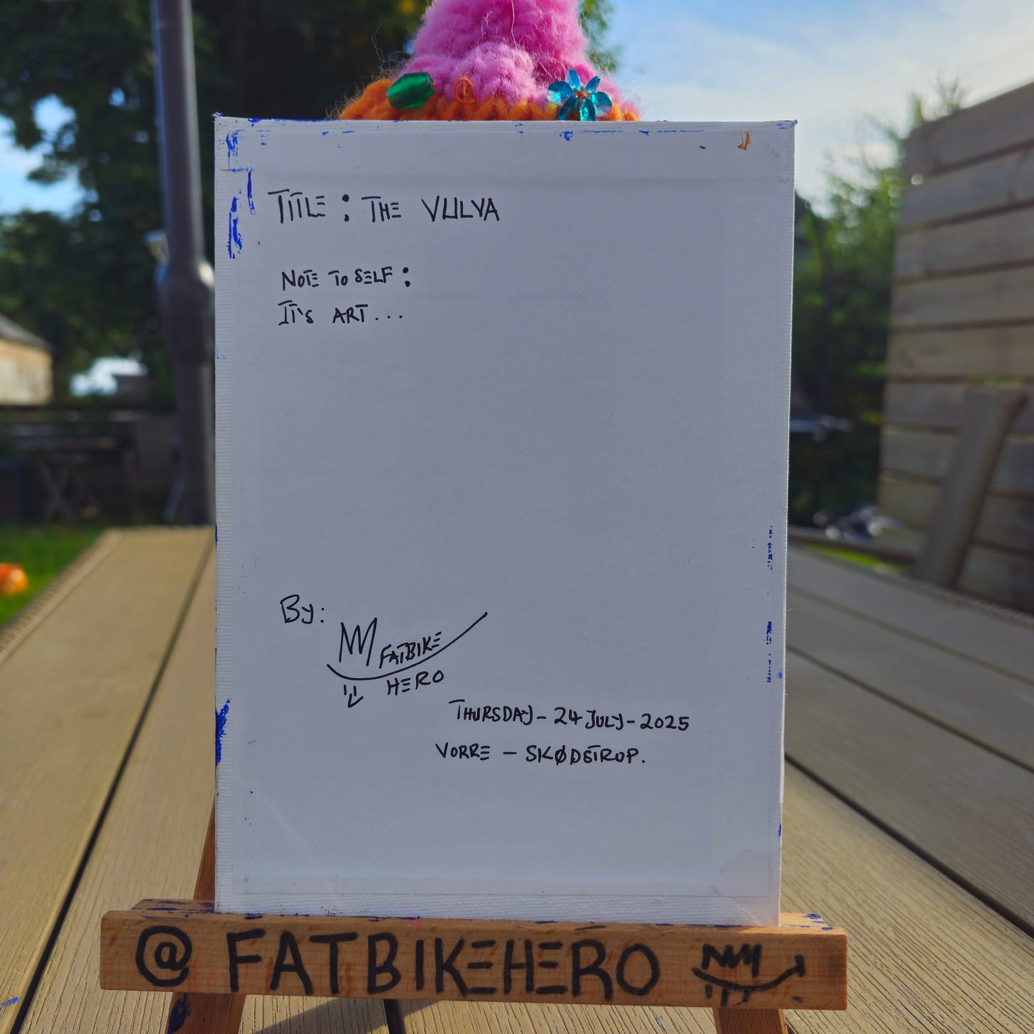 Alt Text: The back of a canvas painting on a wooden easel. Handwritten text in black marker reads "TITLE: THE VULVA", "NOTE TO SELF: IT'S ART...", signed "By M / FATBIKE HERO", dated "Thursday 24 July 2025", location "VORRE - SKØDSTRUP."