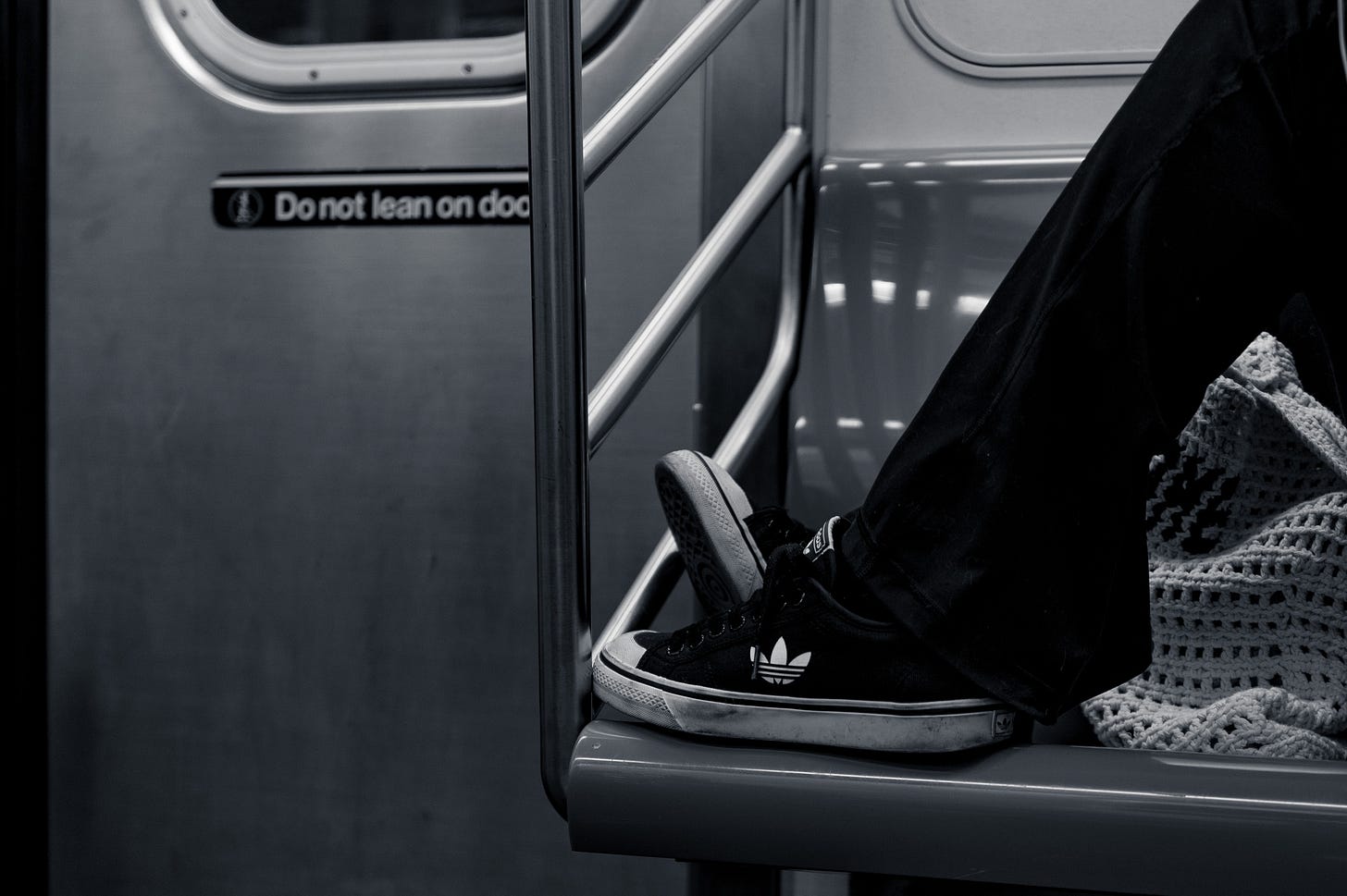 Subway car interior with a person's legs stretched out and Adidas trainers resting on the seat edge beside a crocheted bag, a Do not lean on door sign behind, in black and white.