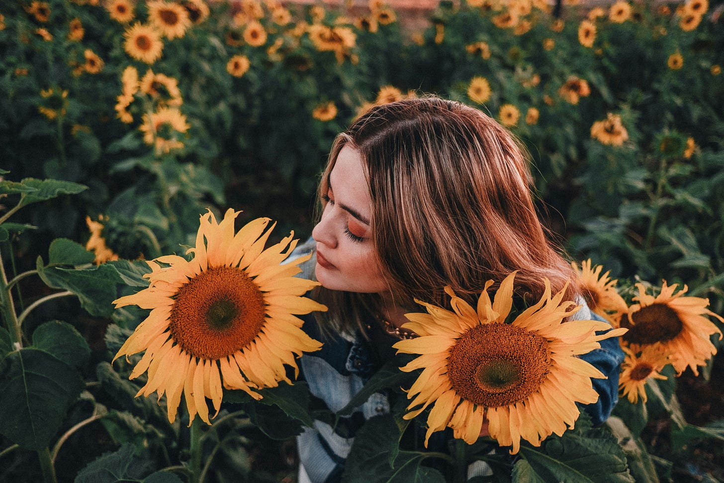 Woma standing in a lush sunflower patch with large vibrant sunflowers on each side of her face, she sniffs one of the flowers on her left. 