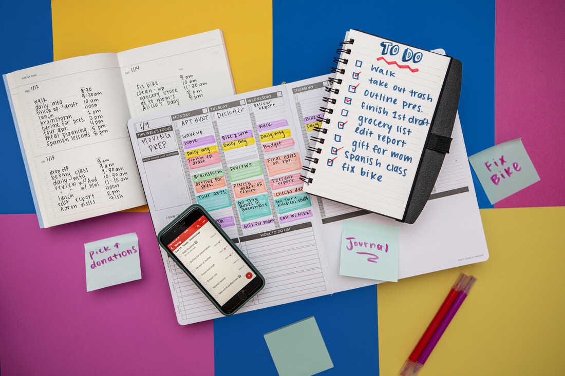 A flat lay photograph of different types of to do lists is photographed from above. Including a planner with time boxing, a to do list in a spiral notebook, a to do list app on a phone and post it notes. The types of to do lists are laid on a background of colorful squares in yellow, fuchsia and yellow.