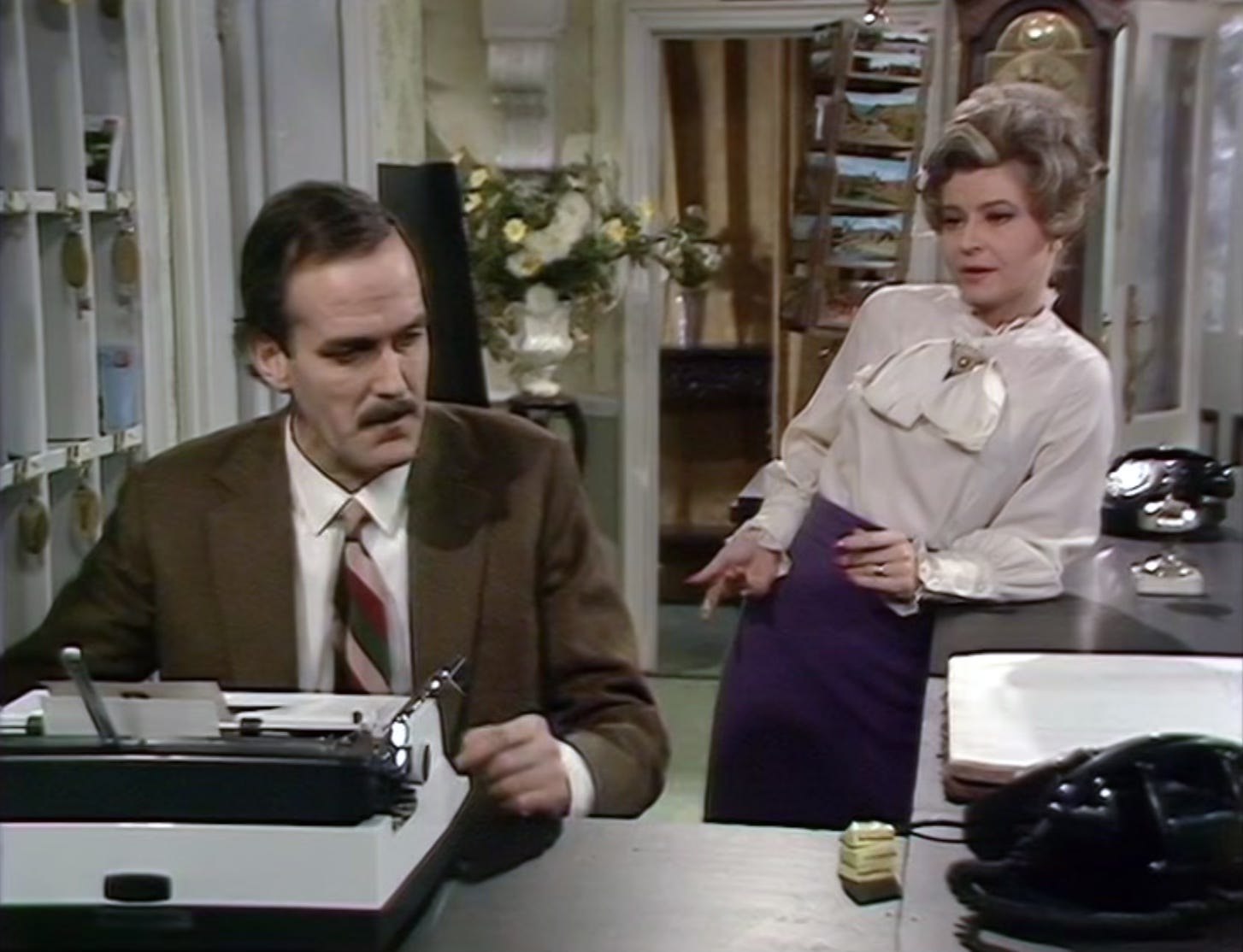 Sybil and Basil Fawlty talking at the reception desk
