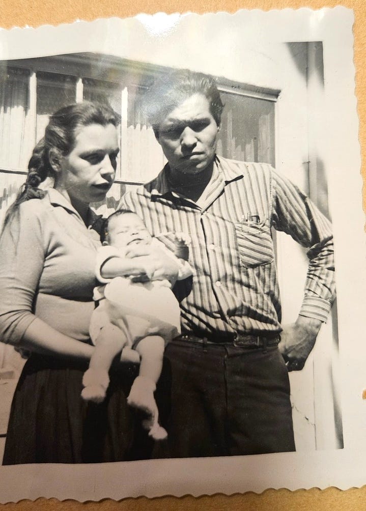 Black and white photo of WInona's mom and dad. Her mom is holding her as an infant in her arms. Her dad is standing next to her mom, in a striped long sleeve collared shirt and dark jeans. Neither is smiling. Winona's mom has brown hair pulled back in a ponytail. 