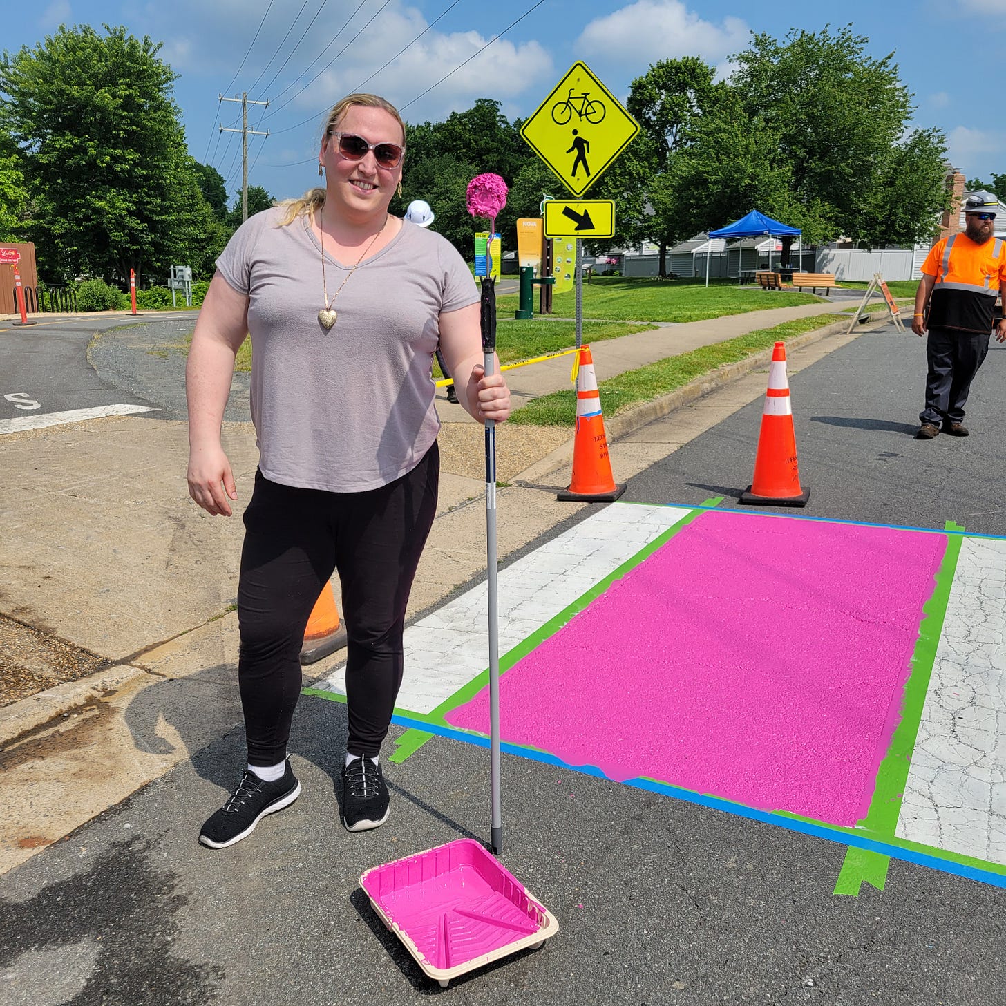 A woman holding a broom stands beside a colorful rainbow crosswalk being painted.