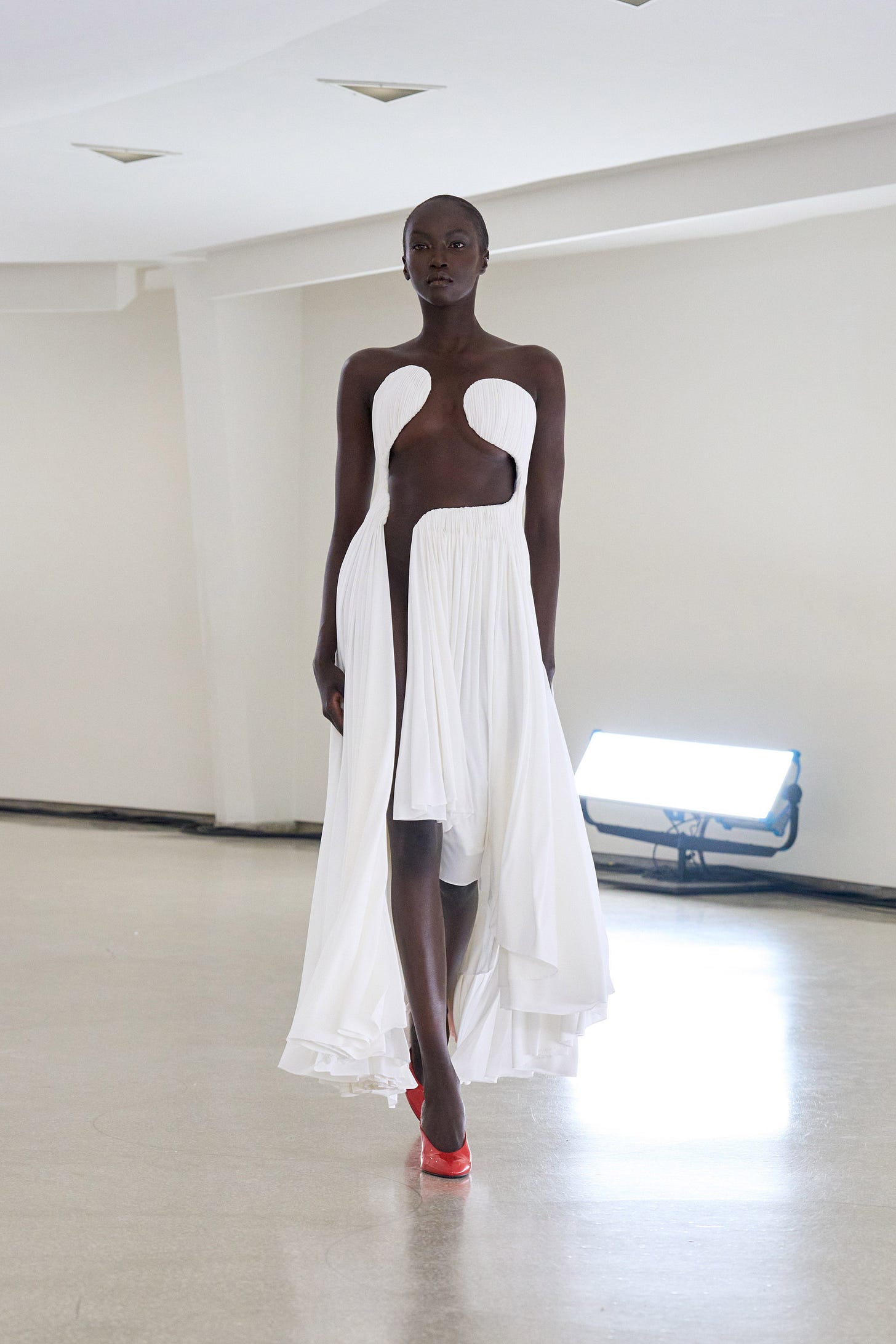 A model walks a curved cement runway while wearing a pleated white dress that has a dramatic cutout running through the front.
