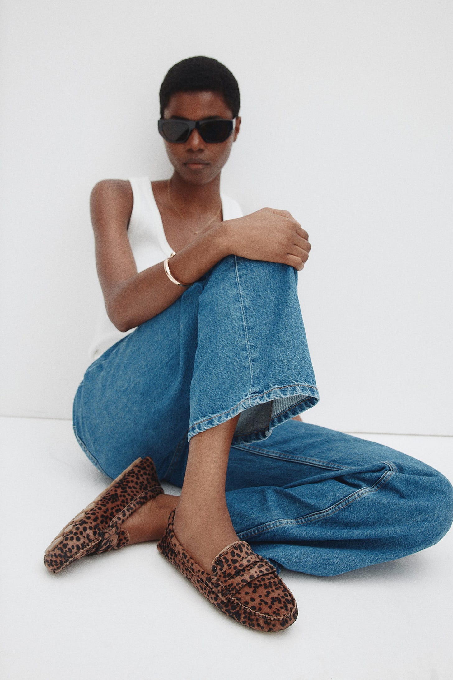 A pair of brown and black leopard print loafers are being worn with blue jeans, showcasing their distinct spotted pattern and classic moccasin construction. One loafer is angled forward, revealing its top and side profile, while the other is angled slightly back.