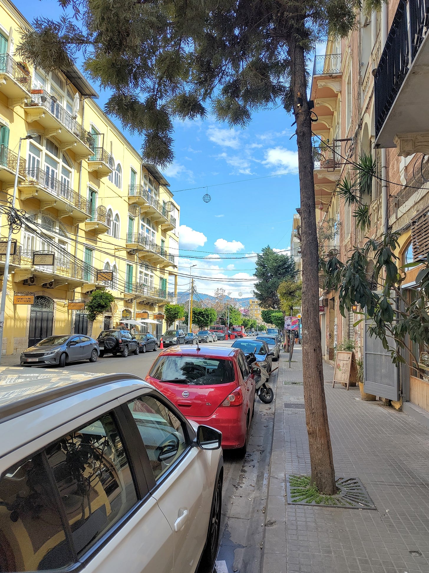 A Beirut neighborhood on a sunny day, with quaint yellow buildings, lush trees, and mountains in the background.