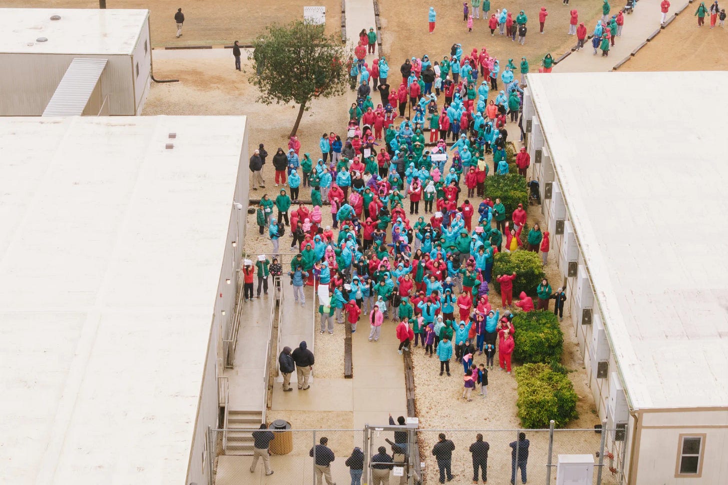 An aerial shot of detainees standing between stark white buildings. Armed guards and a fence hold them in. Several are holding signs. Many appear to be children.