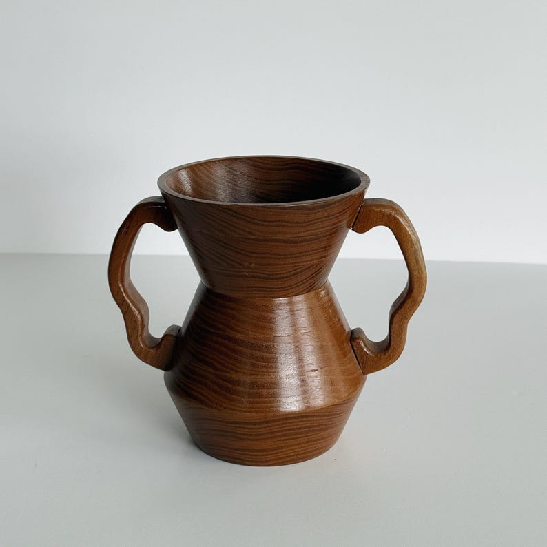 May include: A handcrafted wooden vase with a unique hourglass shape and two curved handles. The vase is made of dark brown wood with a visible grain pattern, giving it a natural and rustic appearance. The design is both functional and decorative.