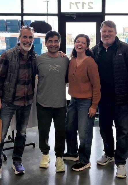 The image shows three people standing together, one in a striped shirt, one in a gray sweater, and one in a blue shirt, all inside a large, industrial-looking building with numbered windows.
AI-generated content may be incorrect.