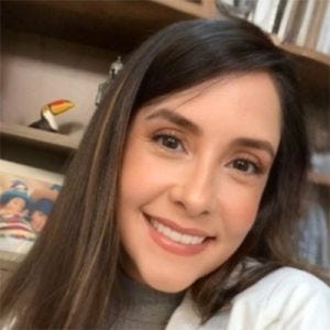 20260820_mariliacristinaoliveirasouza Mulher sorrindo para a câmera, usando blusa de gola alta cinza e casaco branco. Ela tem cabelos longos e castanhos. Ao fundo, uma estante com livros e objetos decorativos.