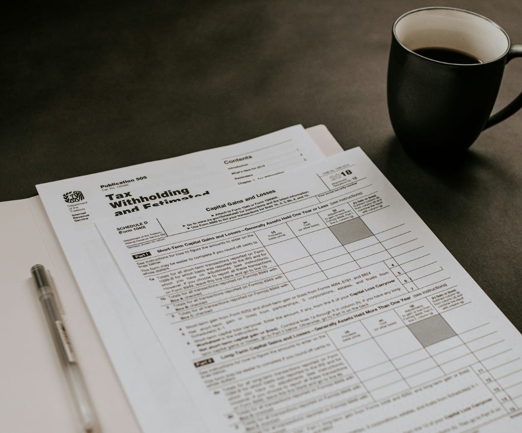 Black and white IRS tax withholding forms are laying on a dark grey desk. There is a pen to the left of the forms and a black coffee mug to the right. The black coffee in the mug is halfway gone, but there’s a light ring on the white inside of the mug where the coffee had been.