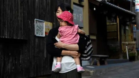 Getty Images A woman in a black cardigan and white shirts holds a baby with a pink t-shirt and a strawberry hat on, in the hot spring town of Arima Onsen on the outskirts of Kobe Getty Images A woman in a black cardigan and white shirts holds a baby with a pink t-shirt and a strawberry hat on, in the hot spring town of Arima Onsen on the outskirts of Kobe