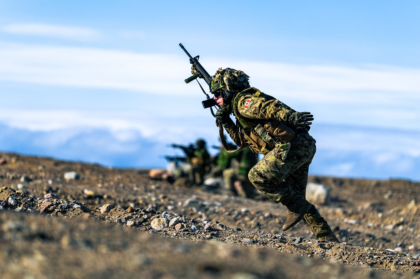 Soldier in camouflage gear sprinting across rocky terrain holding a rifle during a military exercise.