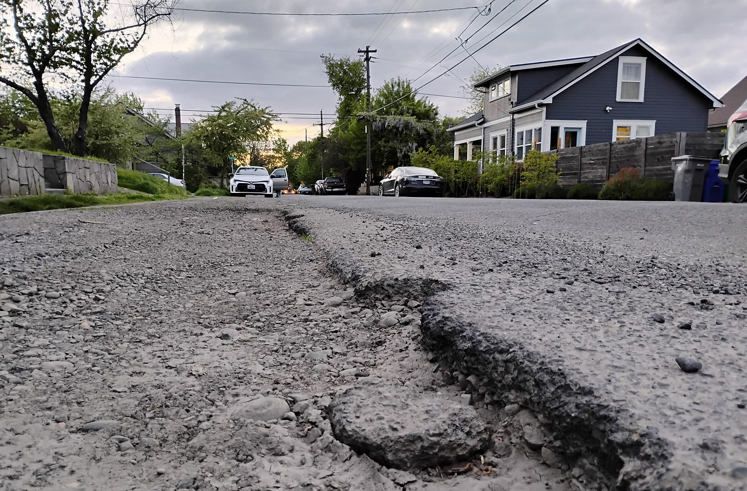 Close-up of a dilapidated road at sunset, with crumbling asphalt in the foreground and a residential area (houses, trees, cars) in the background.