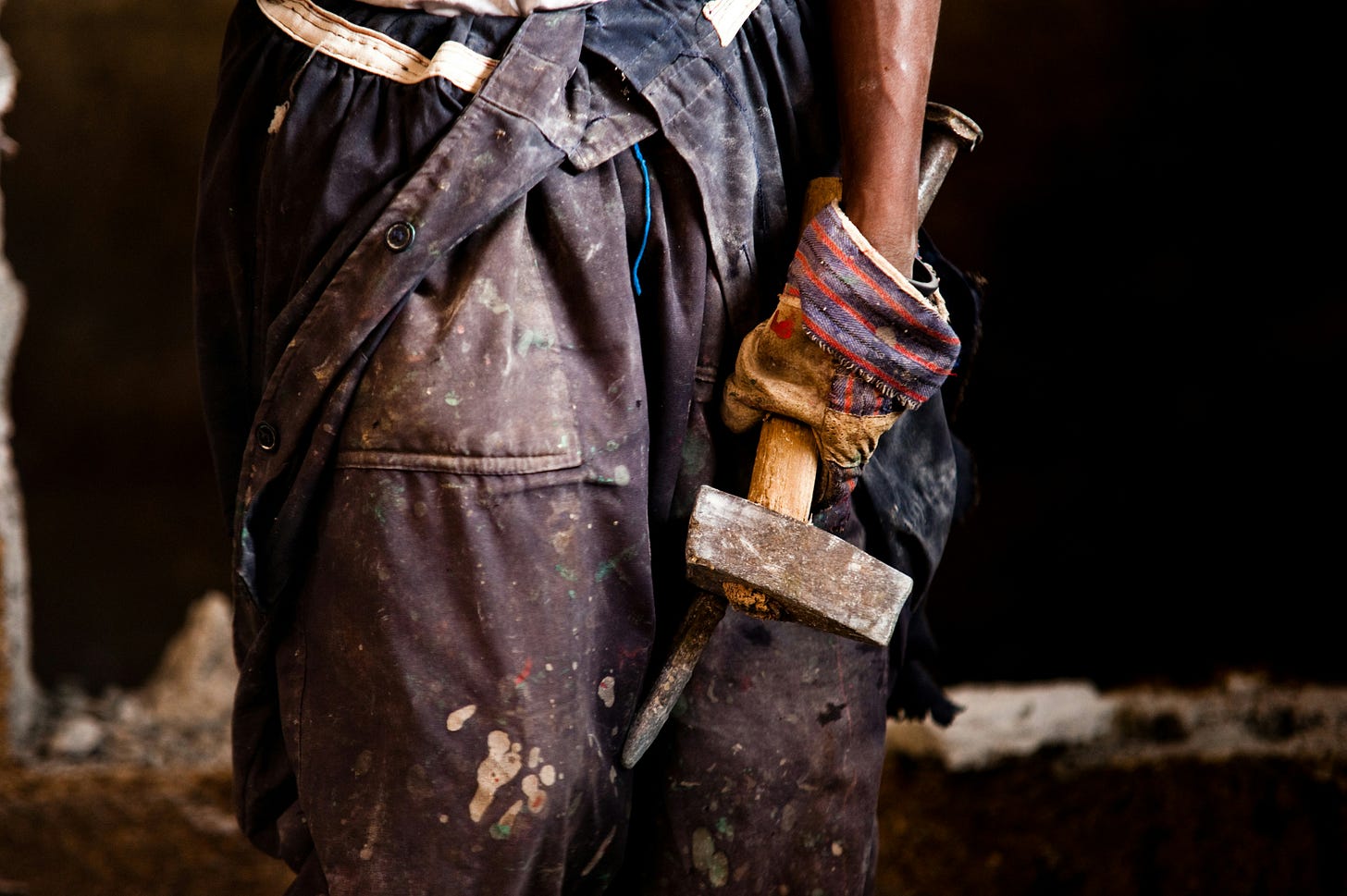 Close-up of a worker's torso and hand, wearing dark, paint-splattered coveralls. A gloved hand grips a mason's trowel at their side, the tool's wooden handle wrapped in worn striped fabric. The background is dark and rough-textured, suggesting a workshop or construction site.