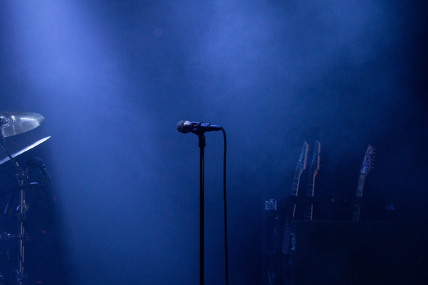 A microphone stand is the focus of this photo. Behind it, an empty drum kit and guitars lie waiting to be picked up and played. The stage is ready, shrouded in smoke, but no one is there yet. 