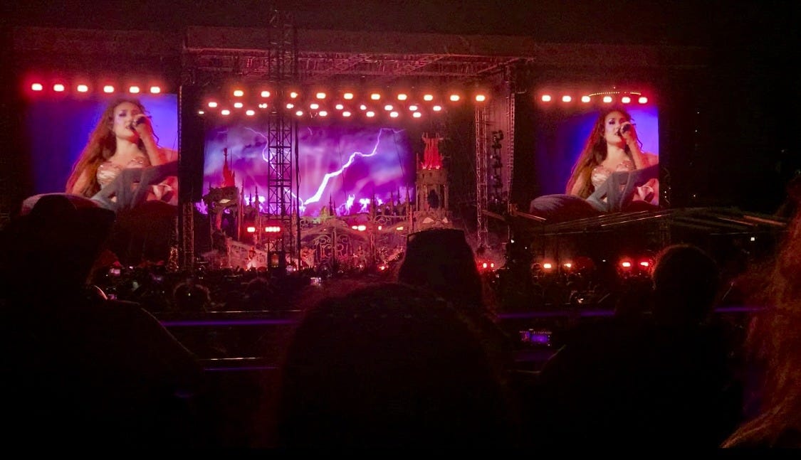 Photo taken from my spot at the Kansas City Chappell Roan concert. Across a sea of heads, an elaborate stage display features a castle-in-a-lightning storm scene. You can just barely make out Chappell Roan singing in the middle of it all. Two giant screens on either side give a closeup shot of her singing into a microphone.